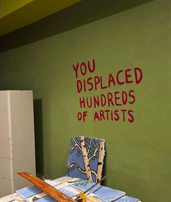 Green wall with red text reading YOU DISPLACED HUNDREDS OF ARTISTS